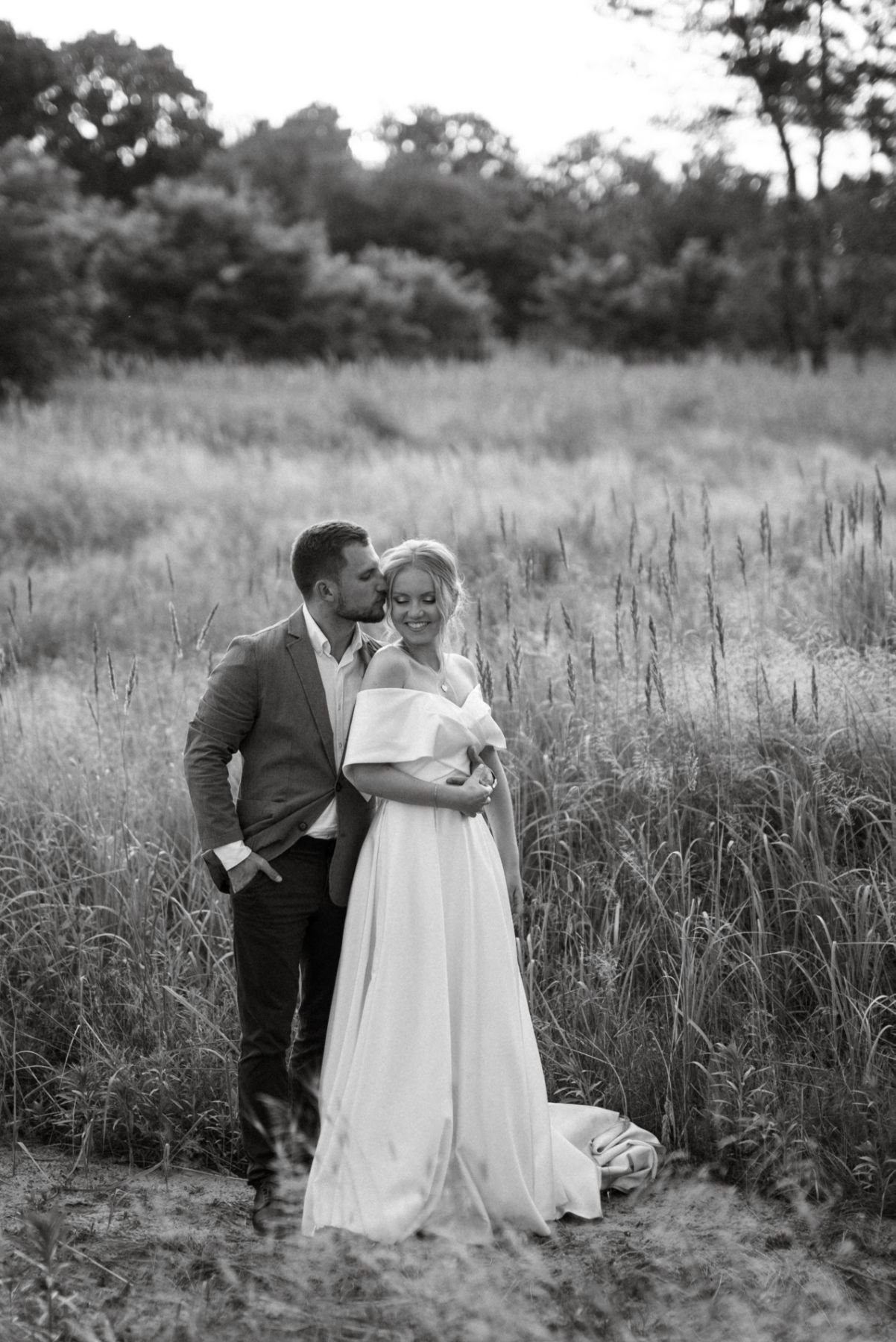 Black-and-white photo of a groom kissing his bride’s temple in tall grass