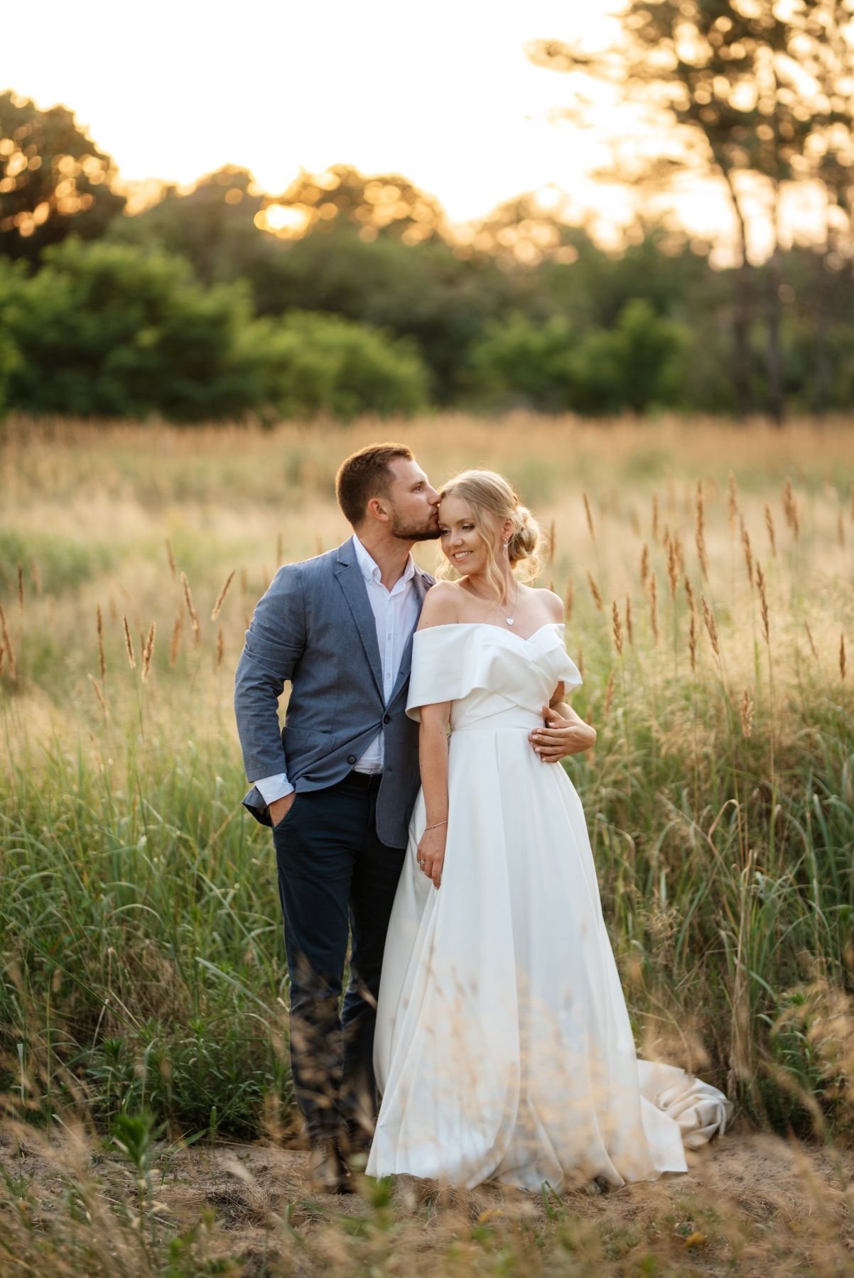 Groom kissing his smiling bride’s head in a golden meadow at last light
